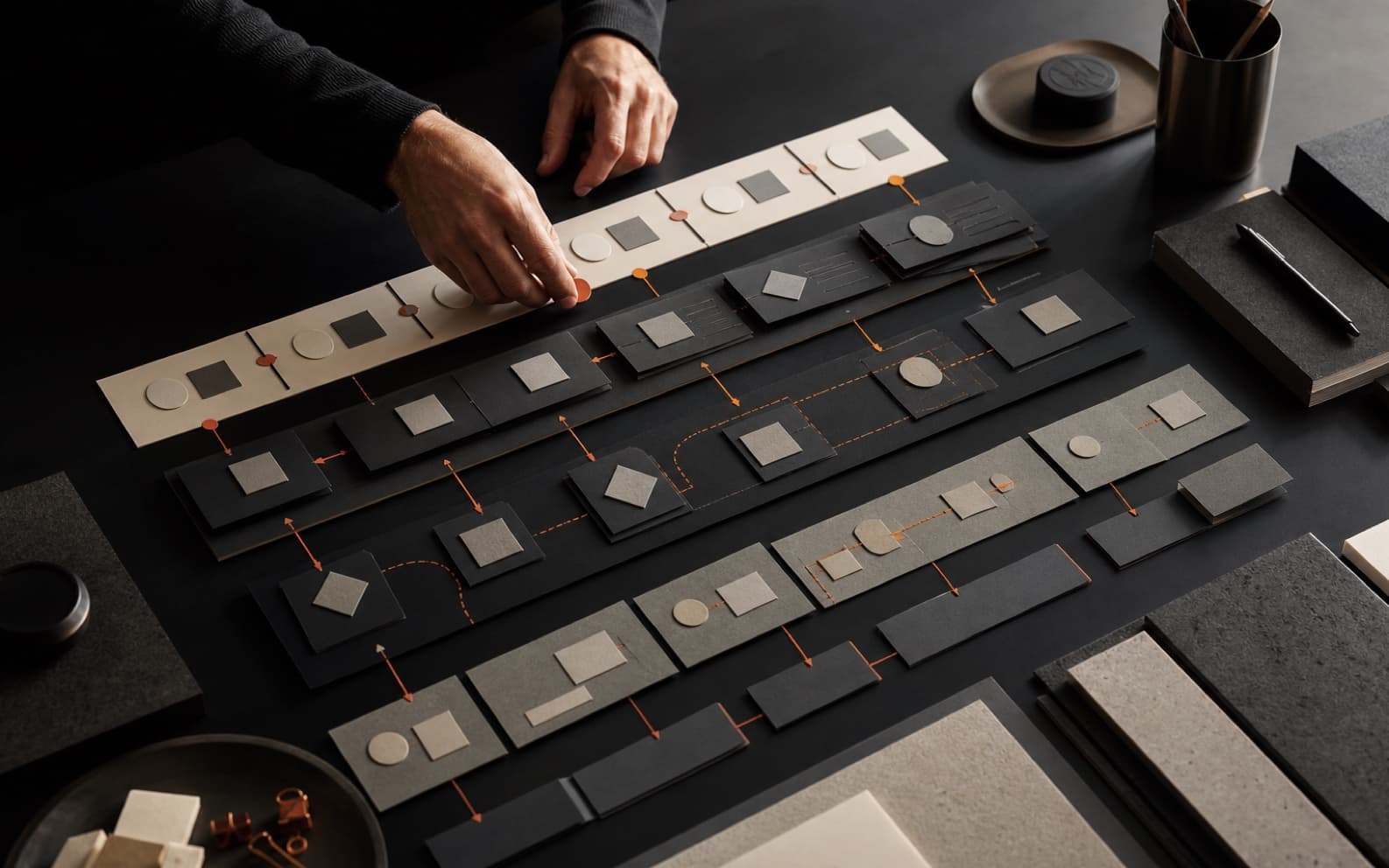 Hands arranging layered off-white and graphite service blueprint cards connected by restrained orange paths on a dark studio table.