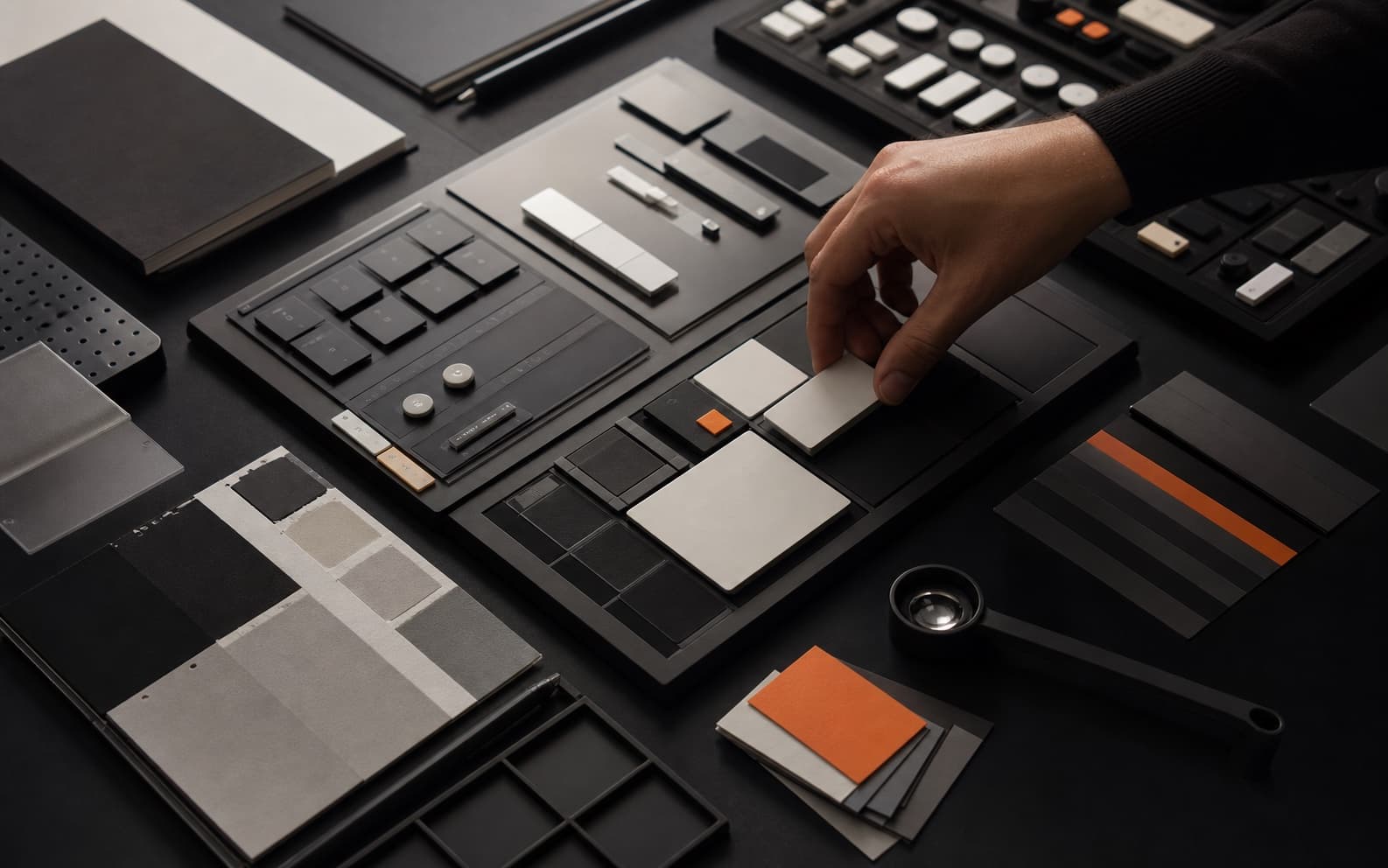 Photorealistic dark design studio table with modular interface tiles, material samples, orange accents, and a hand fitting one reusable component into a shared system.