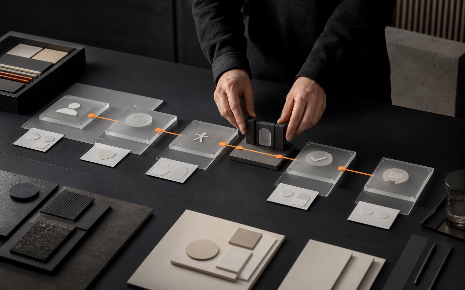 Designer arranging tactile frosted interface modules along an orange path through identity, accessibility, privacy, confirmation, and support stages on a dark studio table.